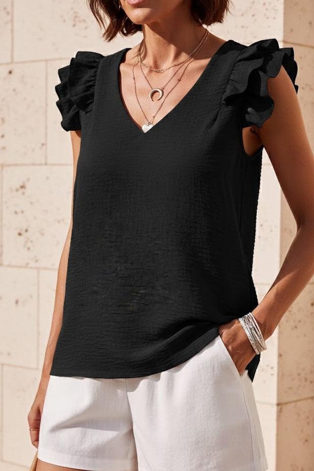 Woman wearing a black sleeveless top with ruffled shoulders and white shorts against a stone wall.